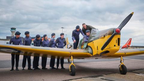 The Miles Magister yellow plane on the forecourt at Luton Airport. A group of eight cadets wearing their purple and black uniform and caps stand behind the left wing, with one sitting in the cockpit and two standing behind him. The pilot wears a green uniform and stands behind the cockpit. The airport control tower is in the background and you can see the orange tail of an easyJet plane.