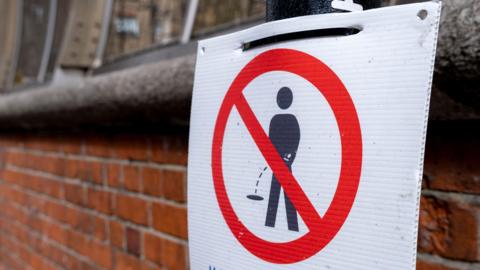 Sign with a person shown urinating with a red cross through it warning urinating in the street can result in a fine