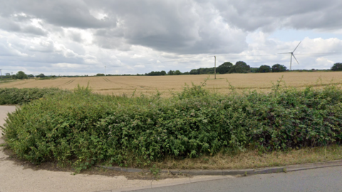 Land off Heath Road in Bagworth, Leicestershire, with shrubs in the forefront at the roadside and trees and a wind turbine in the distance on a dry cloudy day.
