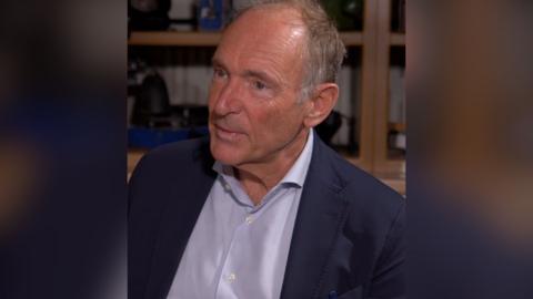 A man, Tim Berners-Lee, wears a light coloured shirt and dark suit jacket. He's sitting down talking to someone off camera.