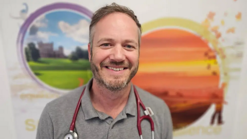 Prof Dominick Shaw from the respiratory clinic is pictured smiling at the camera with a red stethoscope around his shoulders. He is wearing a grey shirt
