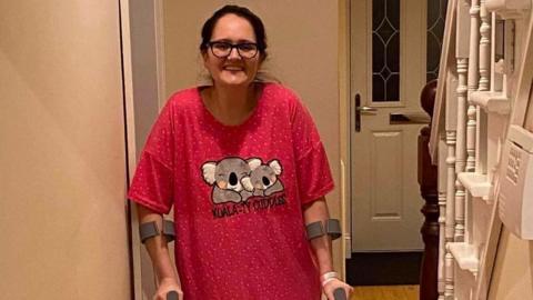 Cat Neill, who is wearing glasses and whose black hair is in a ponytail, stands in a hallway on crutches. She is smiling at the camera.