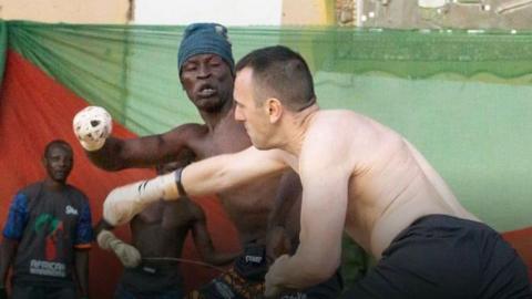 Two topless men, wearing shorts, go to punch one another. Each of one of their fists is wrapped tightly in white rope. There is a Nigerian flag, red and green, behind them on a wall. 