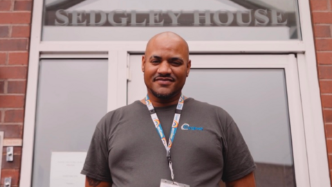 A mid-shot of a bald man standing in front of a doorway with a sign above it that reads "Sedgley House". He is wearing a grey T-shirt and has his hands behind his back. He also has a lanyard with rainbows on it around his neck. 