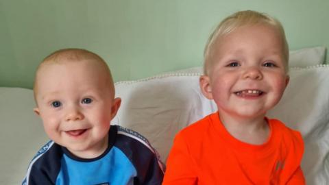 Theodore (left) a nine-month old boy, smiles in a blue top while sitting next to his brother Lincoln, who has a big grin and is wearing an orange top.