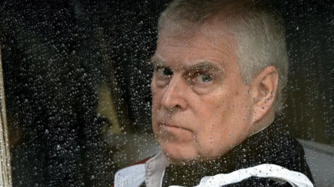 Prince Andrew sat in the back of a Royal carriage with a serious expression on his face. The window is up and you can see droplets of rain on the window.