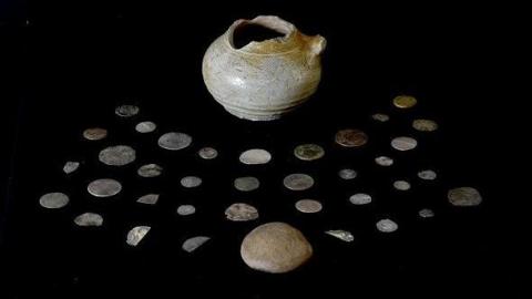 The pot, its lid and the coins are laid out on black fabric. The coins are of different sizes.