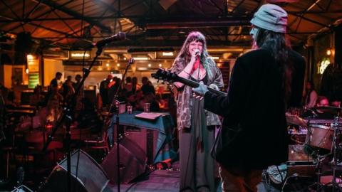 A singer sings into a microphone while a guitarist with their back to the camera plays a guitar. There are instruments on the stage and a crowd of people are standing near the bar in the background. 
