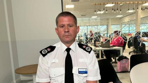 Ivan Balhatchet with short dark hair looking thoughtful and wearing a white police shirt, black tie and black epaulettes. He has a blue and white name badge. An office with people working at laptops is visible behind him.