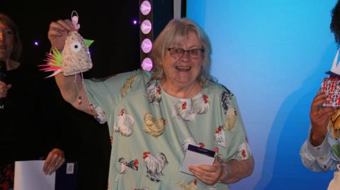 Melody Willers is stood holding a fabric hen aloft at the ceremony as she wears a green shirt covered in paintings of hens.