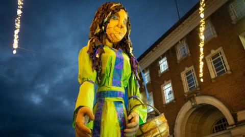 A view looking from the ground at a puppet of a woman with long brown hair and blue eyes. She is wearing a green dress with purple stripes and is carrying a woven basket. Behind the puppet is a brick building and the cloudy night sky.