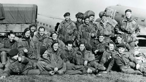 A black and white image of a group of soldiers sitting on the grass in uniform in front of a lorry and an aircraft. 