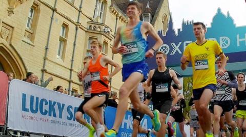 A large pack of runners with advertising boards to either side of them. behind them is a big blue archway saying 'Oxford Half' in white letters.