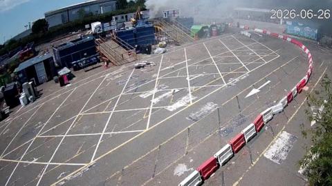 A waste site, with smoke coming from a container, and white and red bollards around it. There are rubbish containers and cabins to the back of the site. 