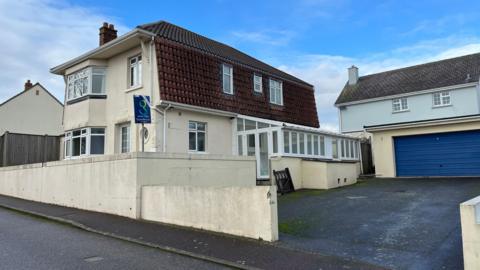 A detached, two storey house with cream walls and an orange, tiled roof. To the right of the image is a separate garage with a blue door. In front of the garage is a tarmac driveway. An estate agent's sign is visible to the left of the image.