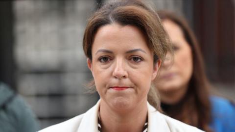 Liz Kimmins has brown hair tied back in a pony-tail. She is wearing a white suit jacket