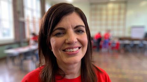 Alana is smiling, she has shoulder-length brown hair and is wearing a red T-shirt.