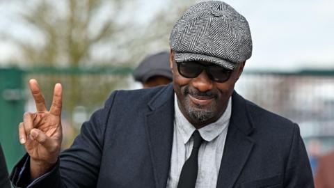 Idris Elba putting up a peace sign with two fingers at Cheltenham Festival while wearing a flatcap and sunglasses