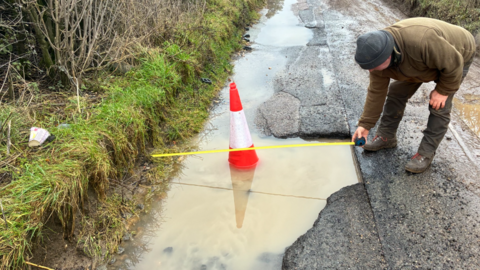 A man holding a measuring tape is bending over a pothole full of rain in Bridewell Lane, Oxfordshire. There is a red and white cone in the water. The single-track road has a grass band on one side. The man is wearing a green jumper and trousers with a grey hat and brown boots.