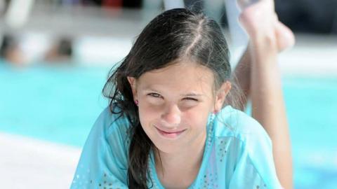 Teenage girl smiling, with long dark hair in a pony tail, lying on her front next to a swimming pool.