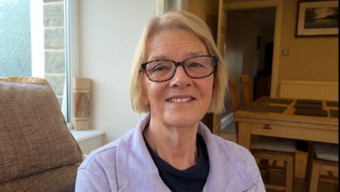 A 72-year old woman with blonde hair and glasses is looking straight ahead and smiling.  She is wearing a lavender coloured jumper with a blue t-shirt underneath.  She is sitting in her conservatory, which backs onto the kitchen.