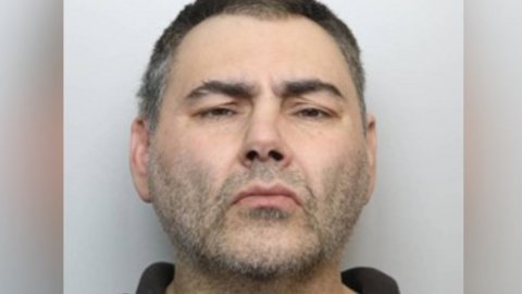 Police mugshot of a man looking directly into the camera. He has a stubbly beard and short dark hair. 