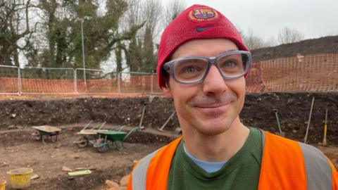 Robert Burns standing in the dig site. He is wearing a red football hat and a hi-vis jacket.