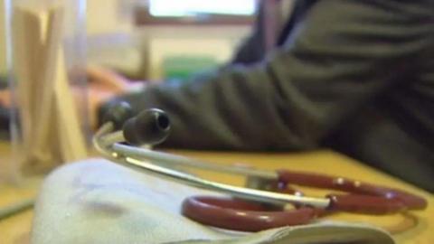 A stethoscope on a GP's desk with the doctor sitting at a yellow desk.