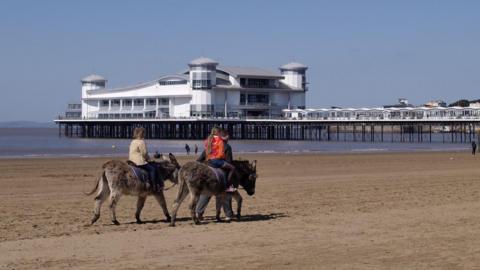 Two people ride donkeys along the beach at Weston-super-Mare. In the background the main structure of the town's grand pier is visible, and the sea is also visible, with the tide about halfway along the pier.