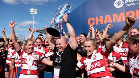 Sean Lynn holds the Premiership Women's Rugby trophy in the air surrounded by the players after winning the 2024 final