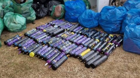 82 purple cylinder-shaped canisters on the grass. They are surrounded by green and blue plastic bags full of rubbish.