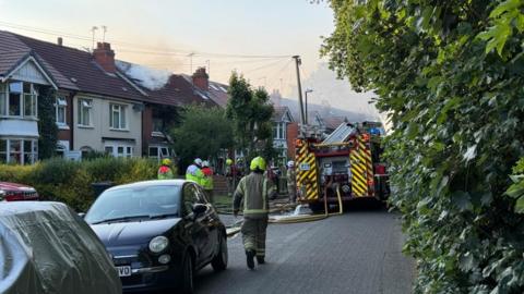 A residential street with a haze of smoke in the sky due a house fire. The street is a narrow lane and has houses on one side and a green bush along the other. Cars are parked in front of the houses and a fire engine is blocking the street. Several firefighters are standing in front of the property and yellow hoses are in the road.