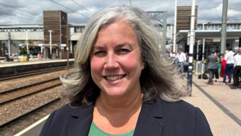 Heidi Alexander wearing a black blazer over a green T-shirt. She is smiling while standing on a train platform.