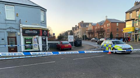 A police car on the corner of a street where a barber shop resides with a police cordon in place.