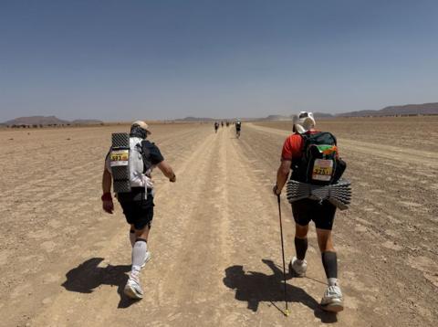 The backs of two men walking through the Sahara desert surrounded by dry land, carrying rucksacks on their backs, one walking with a stick