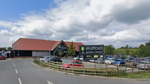 Stratford garden centre. It is a a large building with a red tiled roof. There is a car park in front of it full of cars. Its sign is black with white writing and a green tree logo