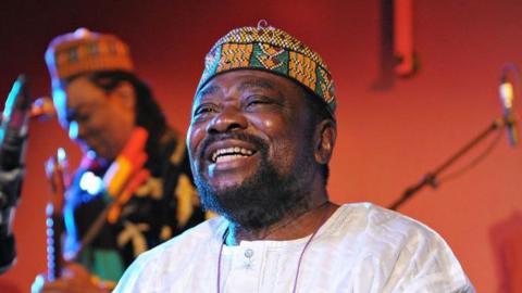 Teddy Osei, dressed in a crisp white linen shirt and a traditional African cap, radiates joy with a broad smile as he performs with his band Osibisa at the Front Room of the Queen Elizabeth Hall in 2010.