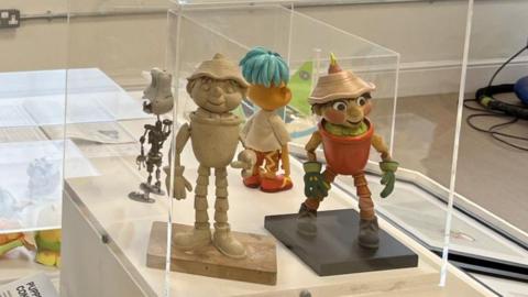 Plasticene figure of a flowerpot man with hat and terracotta pot top next to a painted version in a display case at the exhibit.