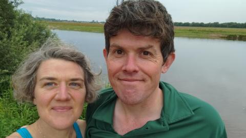Nell and her husband smiling together in front of a lake