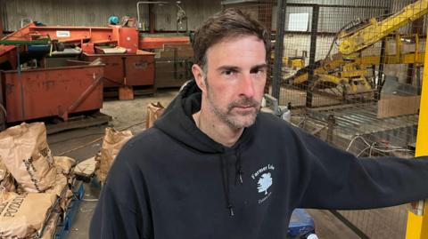 Luke Abblitt has short brown hair swept across his head. He has a stubbly, greyish/dark beard and moustache and is wearing a black hoodie, with the hood down. Behind him are brown paper sacks of potatoes and machinery to bag them.