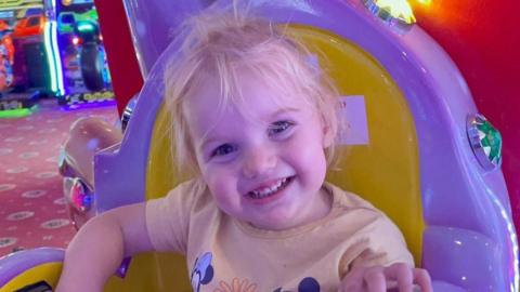 A young blonde toddler wearing a yellow t shirt smiling into the camera. She is sitting on a yellow and purple chair at an arcade. 