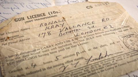 A piece of paper with the words gun licence at the top of it.  The name Ronald Kray are written in by hand and his address is given as 178 Vallance Road, Bethnal Green, London, E2.
It also has an official  date stamp of 30 May visible to the right and the signature of the official who issued the licence.
It was issued at '4 hours 5 minutes pm'.