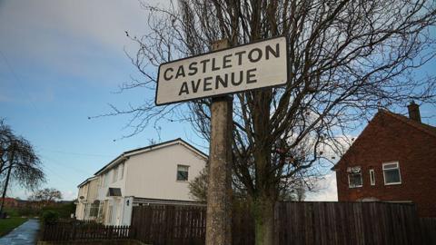 A street sign saying Castleton Avenue with white houses and a fence and trees in the background.
