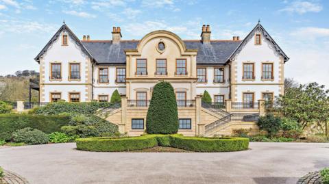 The entrance to a mansion. The property is a beige and white colour with perfectly trimmed bushes in front of the house. A circular driveway can be seen looping around the entrance to the property.