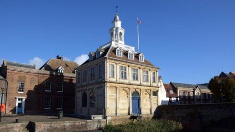 The Custom House, a two-storey light brick building with windows jutting out of the roof. On top of the roof is a spire-type tower.