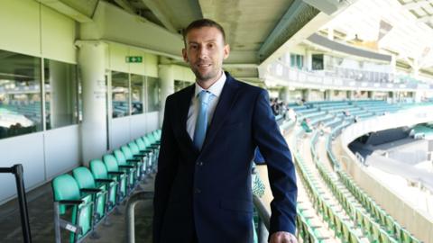 Chris Head, wearing a navy suit and light blue tie. He has short dark hair and stubble. He is standing next to rows of seats for spectators at a cricket ground.