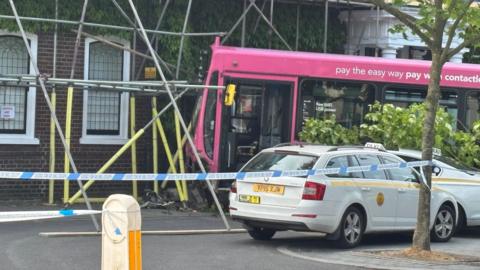 A close up view of a bus that has crashed into a building. The front of the bus has been damaged. The building has scaffolding on it which has been destroyed by the bus. A police cordon can be seen. Two taxis caught up in the collision rest next to the bus.