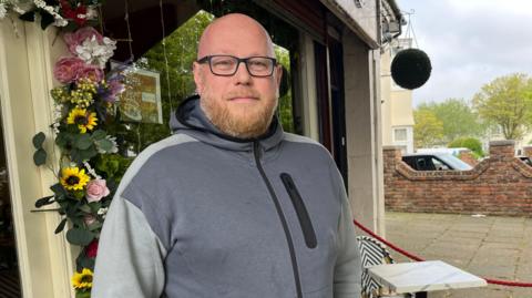 James stands for the photo outside his coffee shop, near to the proposed plans for new cycle infrastructure. He is wearing a grey hoodie, is bald and has black glasses.