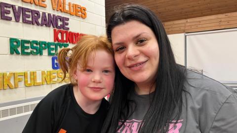 A mother and daughter are smiling. The young girl on the left has ginger hair in pig tails and is wearing a black T-shirt. The mother on the right has long dark hair and is wearing a grey T-shirt. Behind them there is writing on the wall which includes the words kindness, respect and thankfulness.