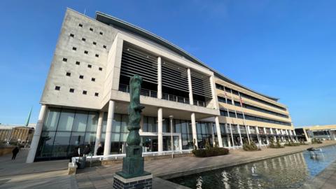 The Harlow Council building is a white brutalist concrete design, with pillars propping up the first one or two floors. There is a sculpture outside and a water feature. There are blue skies above.
